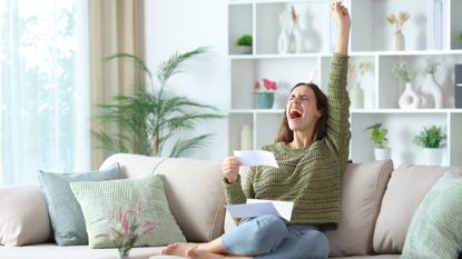 Excited woman celebrating with her fist in the air while holding a bill and sitting on the couch at home