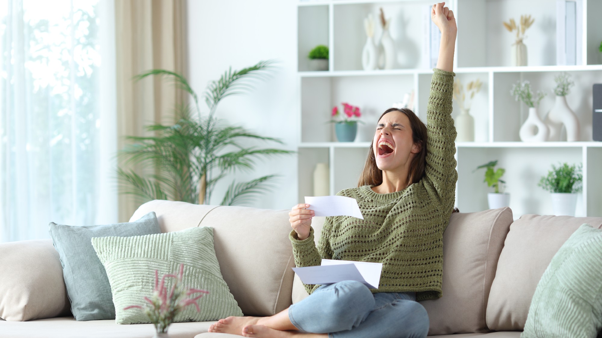 Excited woman celebrating with her fist in the air while holding a bill and sitting on the couch at home