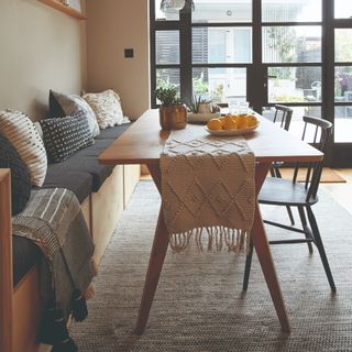 A dining room with a textured table runner, a rug beneath the dining table and a throw with cushions arranged on the banquette seating