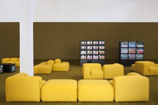 A waiting area room with yellow sofas/chairs. Against the wall are two blocks of four-by-four televisions and three-by-three televisions.