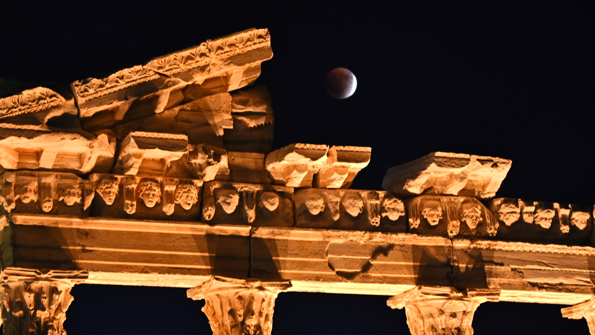 A mostly eclipsed full moon is pictured hanging above the collapsed roof of an ancient temple which is lit by yellow light. The moon's right crescent is lit, with the rest bathed in deeper shadow.