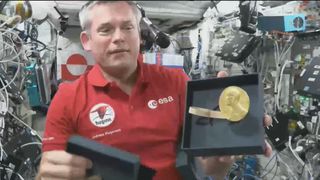 An astronaut in a red shirt holds a gold Nobel Prize medal in space