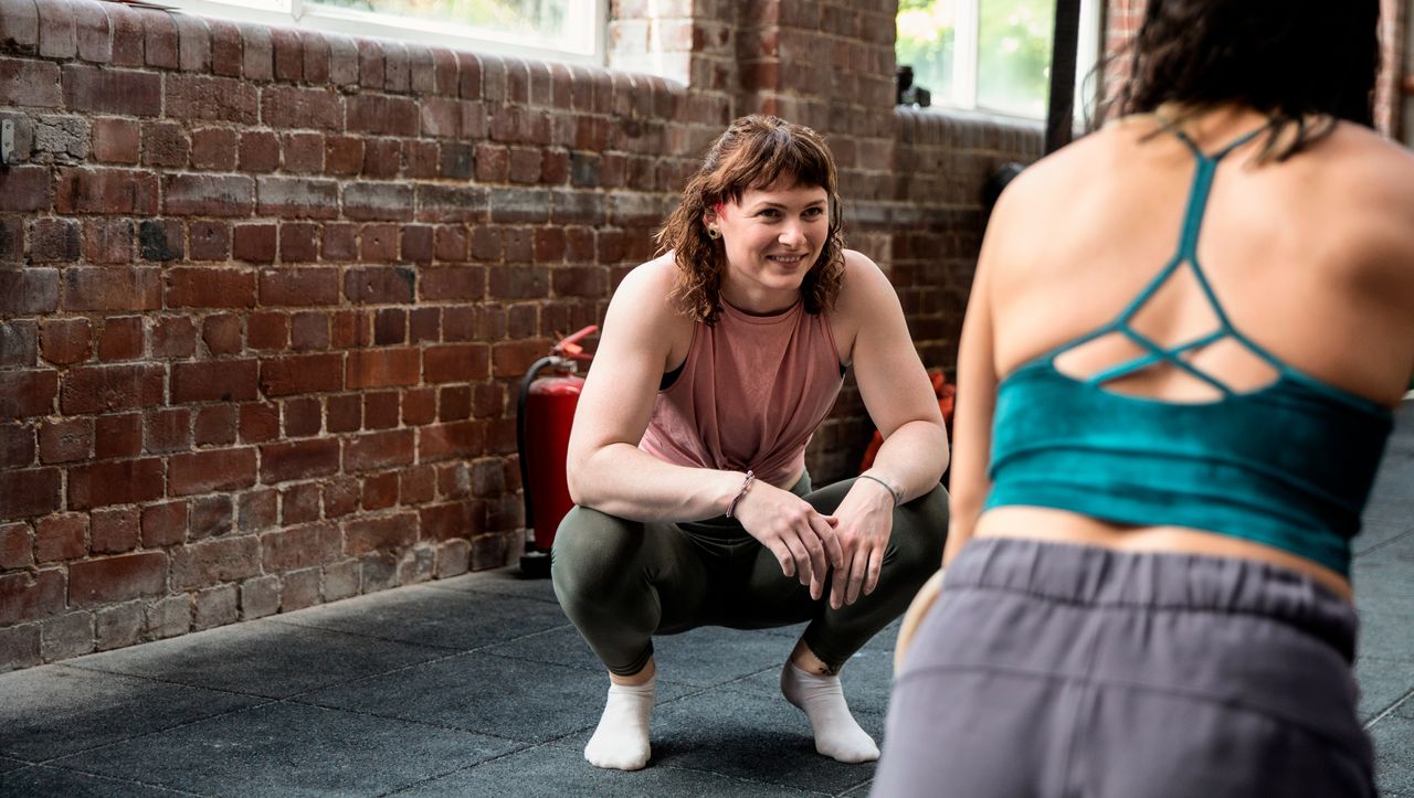 Woman in gym crouches and looks at a woman with her back to the viewer.