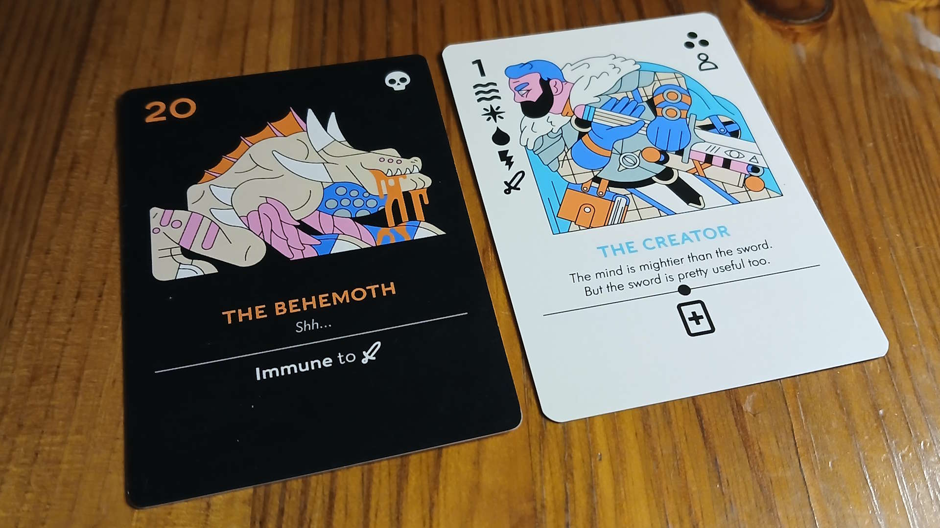 A card showing &#039;The Behemoth&#039; faces a &#039;The Creator&#039; card on a wooden table
