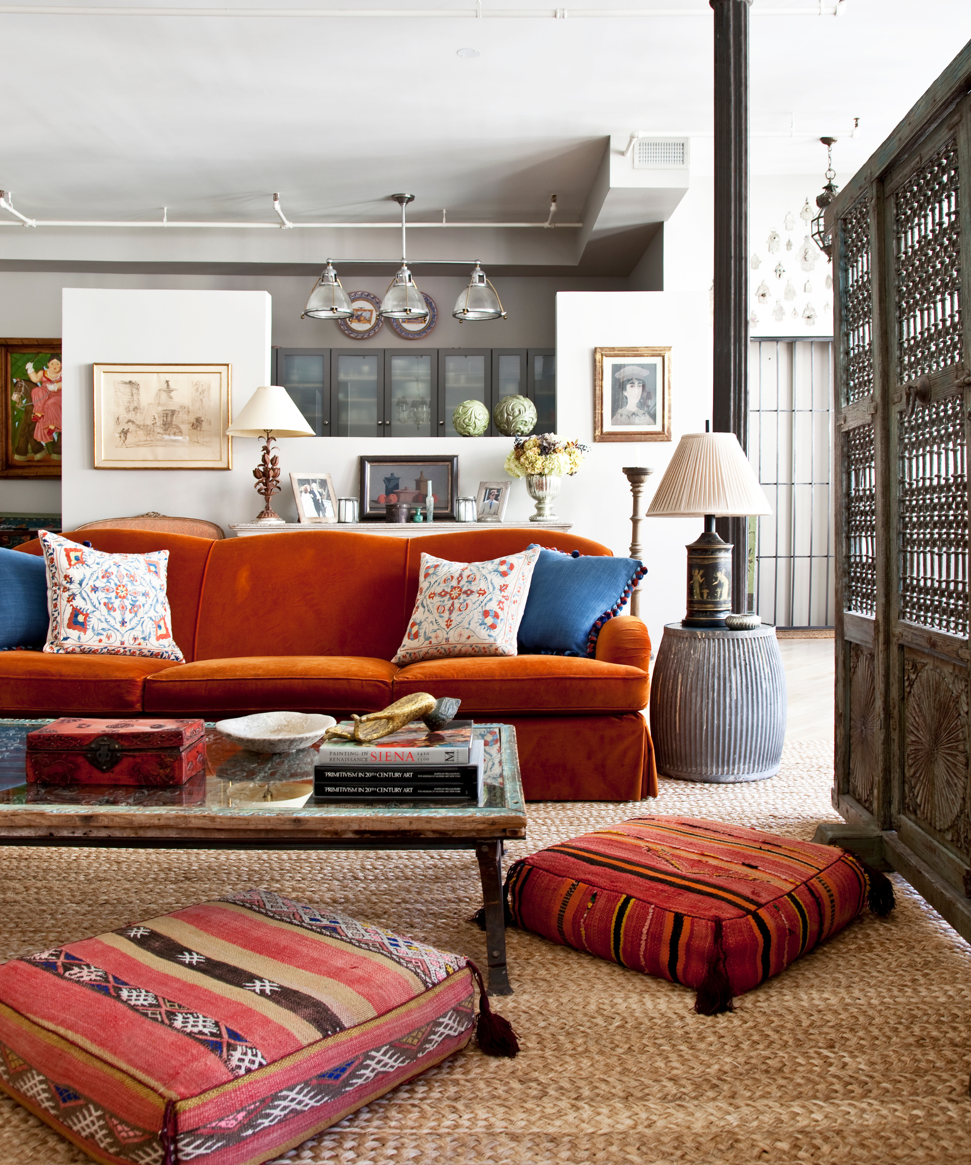 a living room with a vibrant orange couch, two floor pillows, a jute rug, and a coffee table with books and decorative objects on top