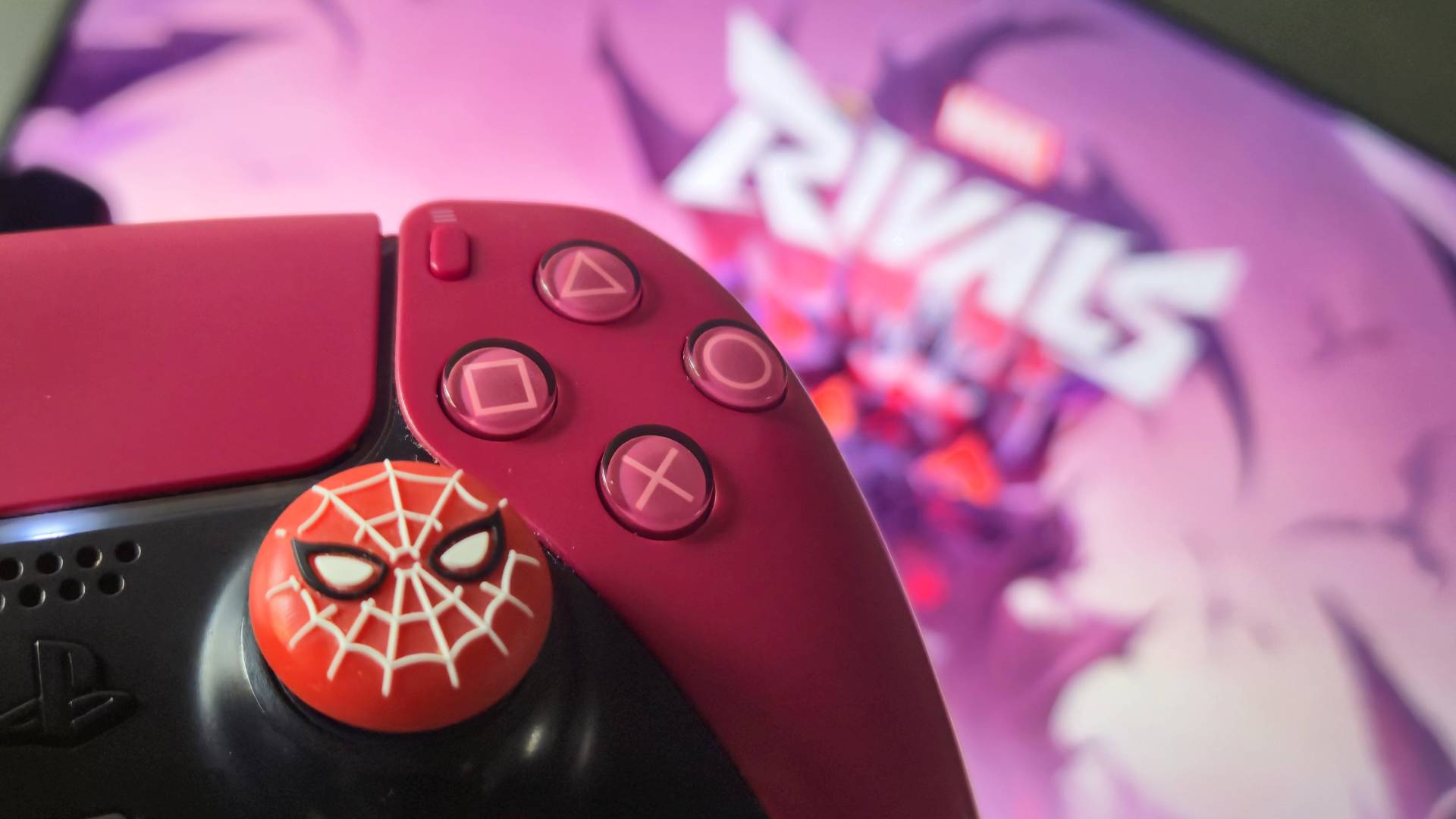 Photo by Rosalie Newcombe of a red DualSense with Spider-man stick grips in the foreground, and Marvel Rivals playing on the TV in the background.