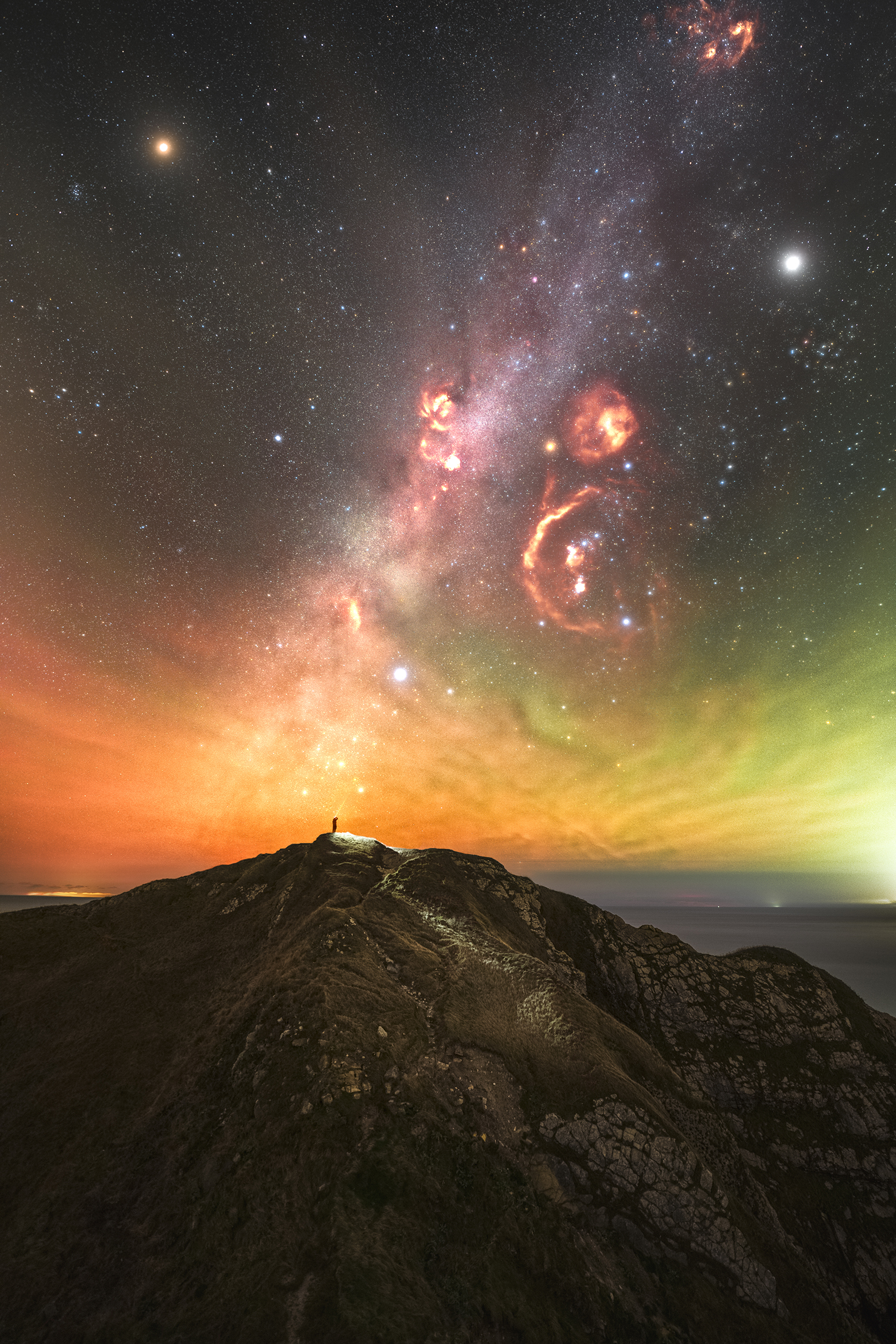 A vibrant night sky filled with stars and colorful nebulae, above a mountain peak silhouetted against an orange horizon