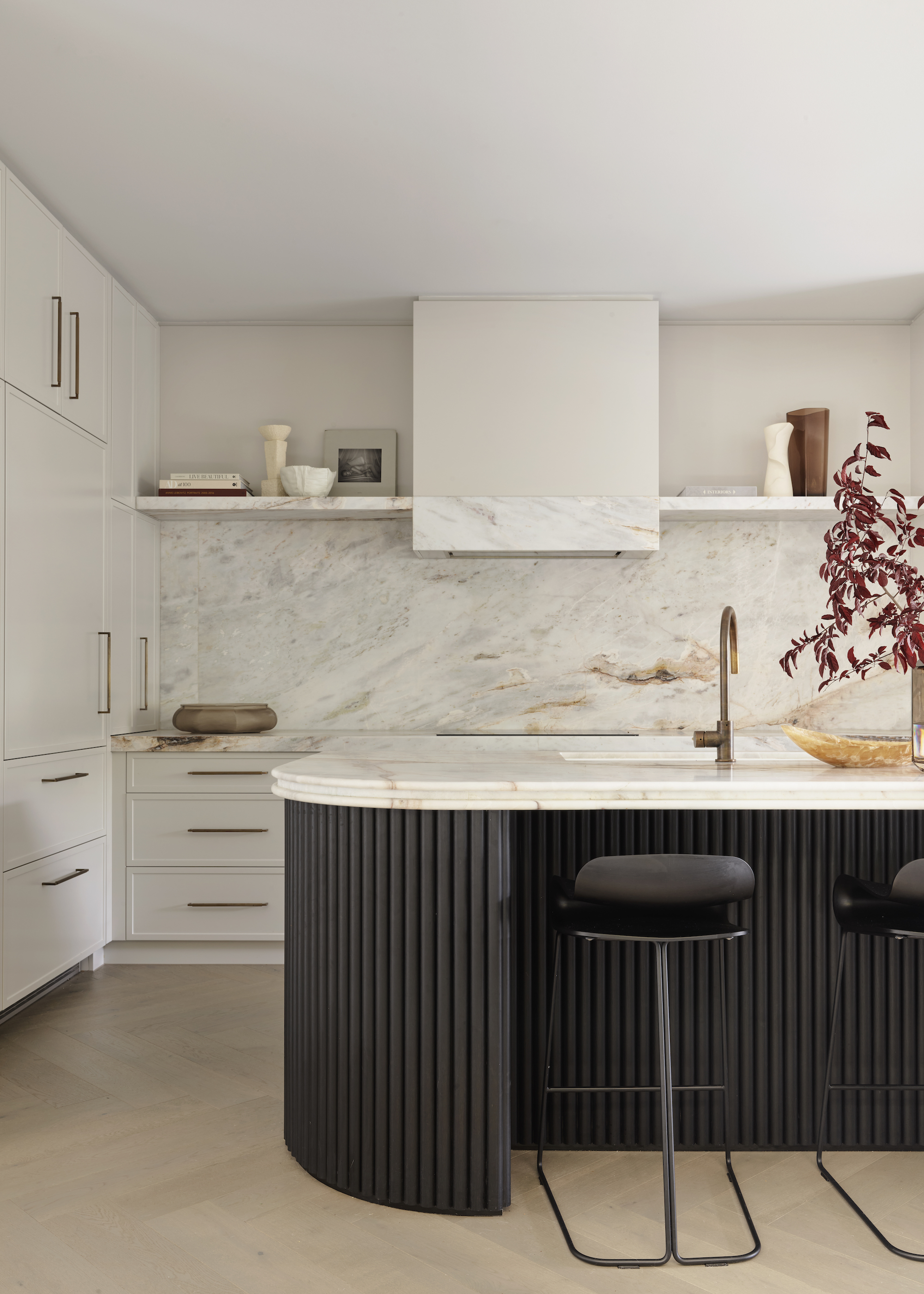 marble island with dark base and black stools. On the island is a sink and tap as well as a large plant.