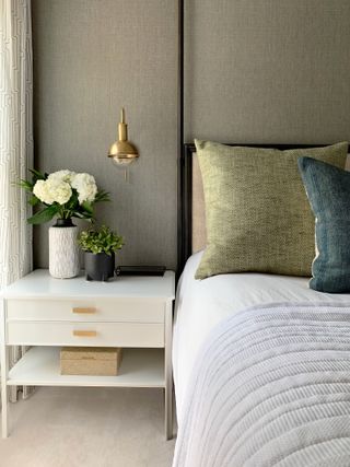 olive green textured wallpaper in bedroom with green textured cushion, blue textured cushion, white side table, gold wall light, black metal bed, white bedding