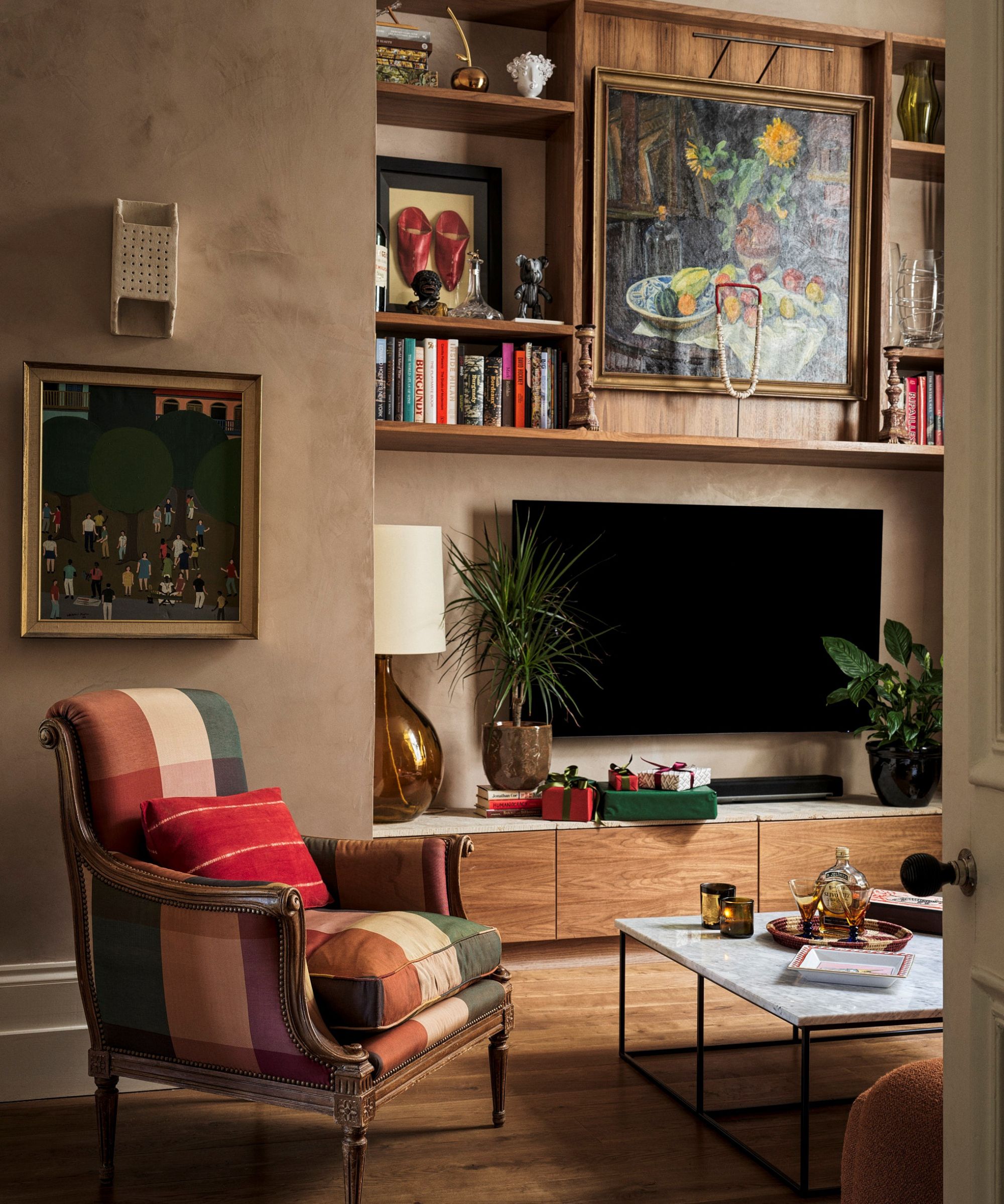 Living room with brown plaster walls and an alcove with built in shelving with a TV below and shelves with characterful decor above. An armchair in purple, green and cream check sits next to a coffee table with a marble top
