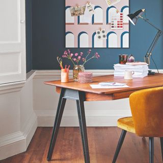 A navy-painted home office with a candle on the desk and a velvet yellow chair paired with it