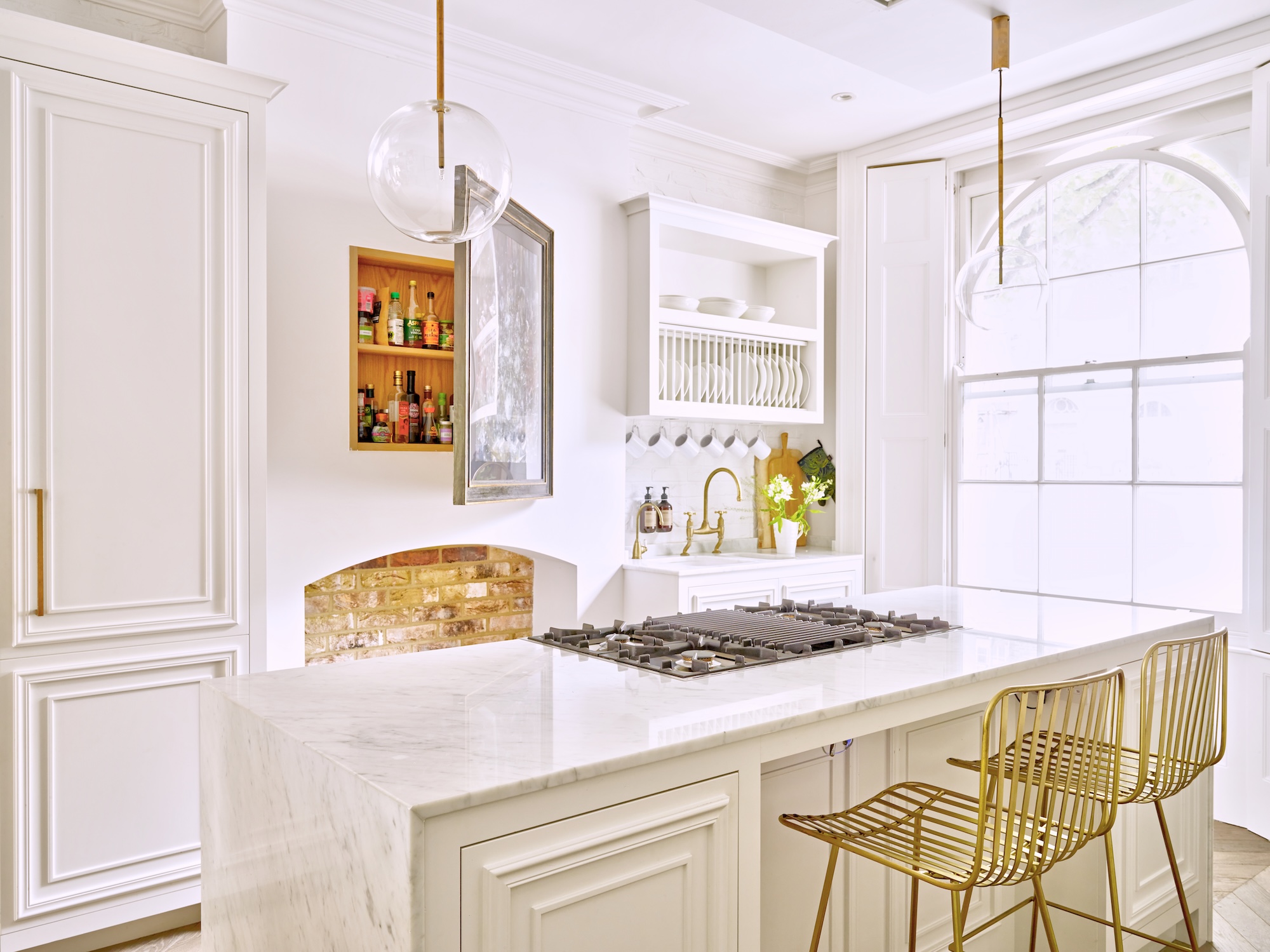 An all white kitchen with a fireplace, oversized arch window and island with two bar stools and hidden wall storage