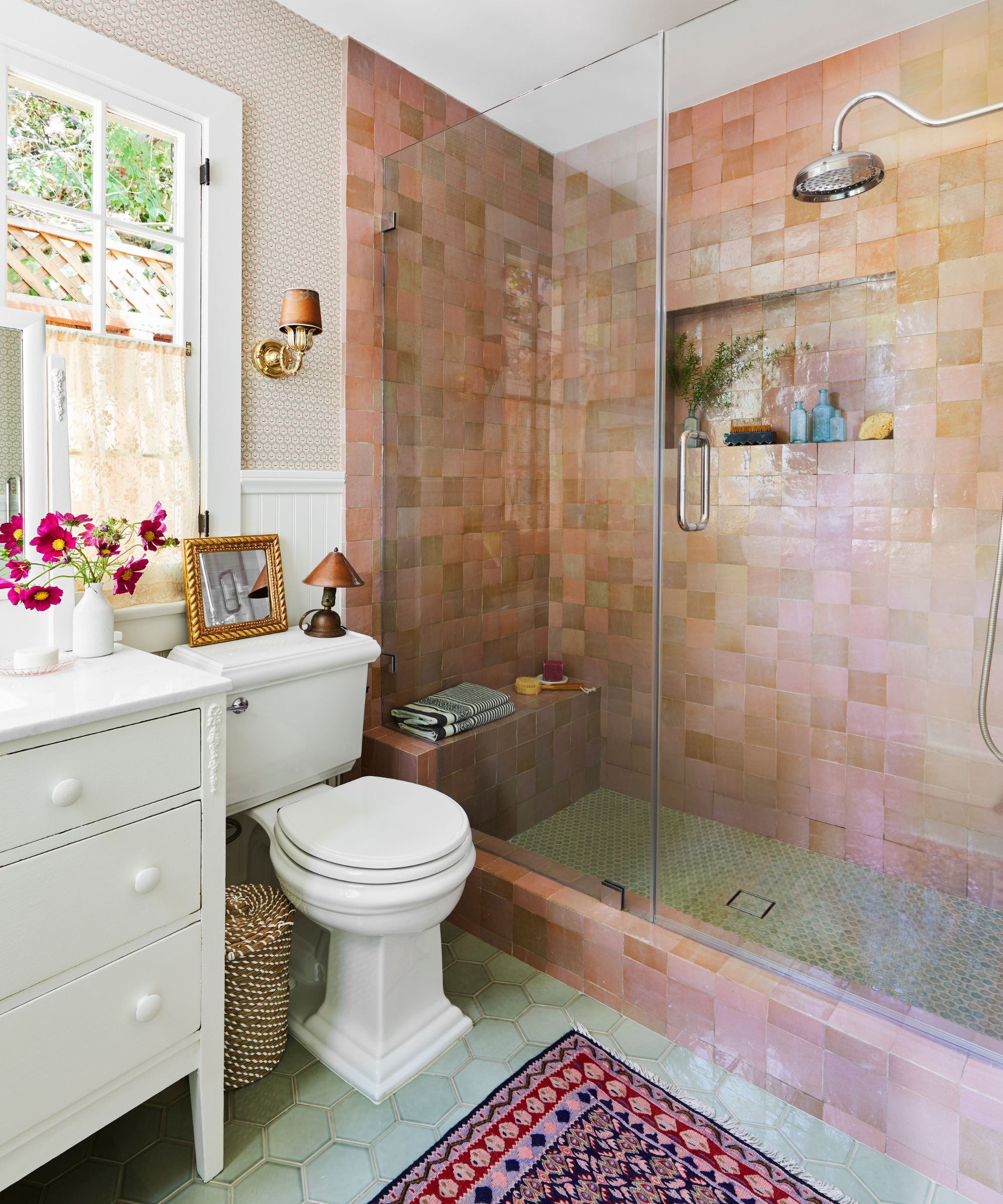 A modern rustic bathroom with muted pink zellige tiles in the shower, light green hexagonal floor tiles, a dark pink and electric blue patterned rug, and a white vintage vanity unit by the window.