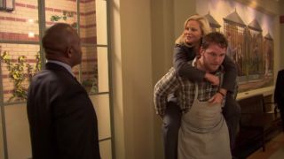 Amy Poehler and Chris Pratt as Leslie and Andy, doing a piggyback race through city hall