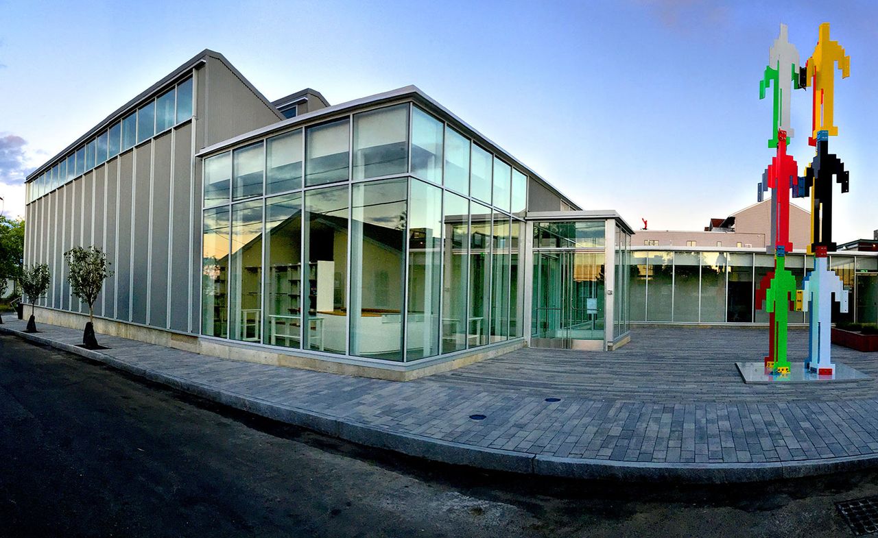 A building with large glass windows, planted trees and an exhibit made of different coloured men shaped objects on top of each other in front of it.