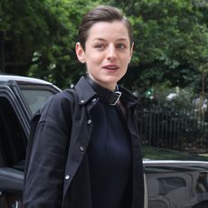 Emma Corrin wears a black button-down shirt over a black dress