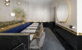 Dining room with grey and blue furniture, black marble and brass highlights on the walls, and a large circular mirror