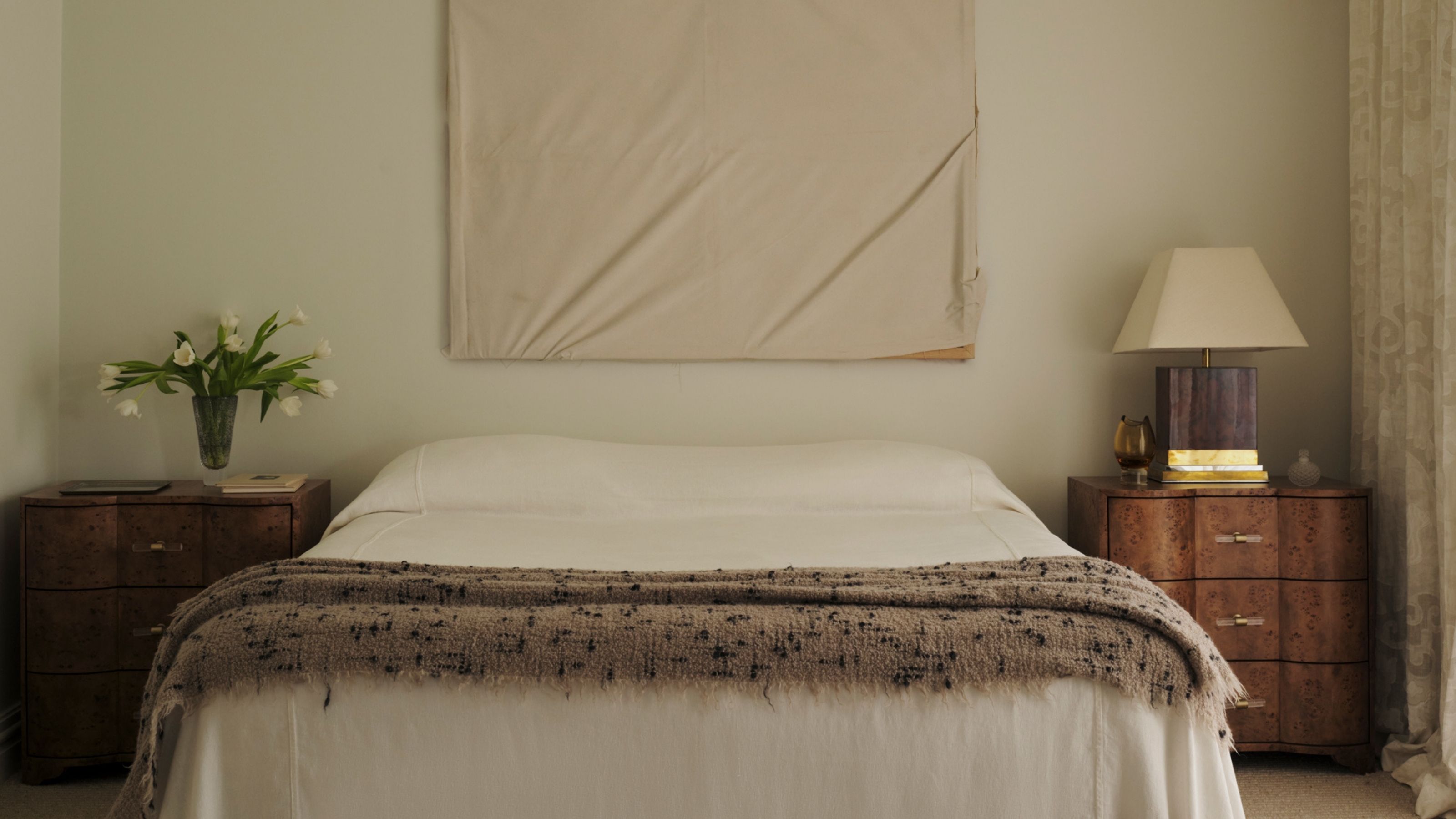 Image of a minimalist bedroom with textured details. There is a white bedding with a beige, textured throw, burl wood nightstands with a lamp and a vase of tulips.