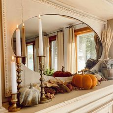 A fireplace mantel with oval mirror, pumpkins, pampas grass in a vase, candlesticks, pine cones and fall foliage