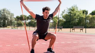 Abs and arms workout: Man performing a squat and overhead press outdoors on athletics track during resistance band workout