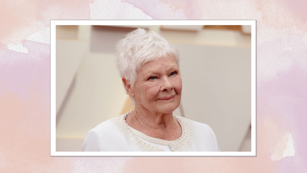 Judi Dench smiling with a grey pixie cut and wearing an embellished white jacket, on a pink and purple watercolour background