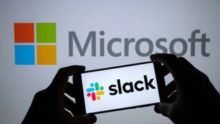 A person holding a smartphone with a Slack logo against a larger Microsoft logo