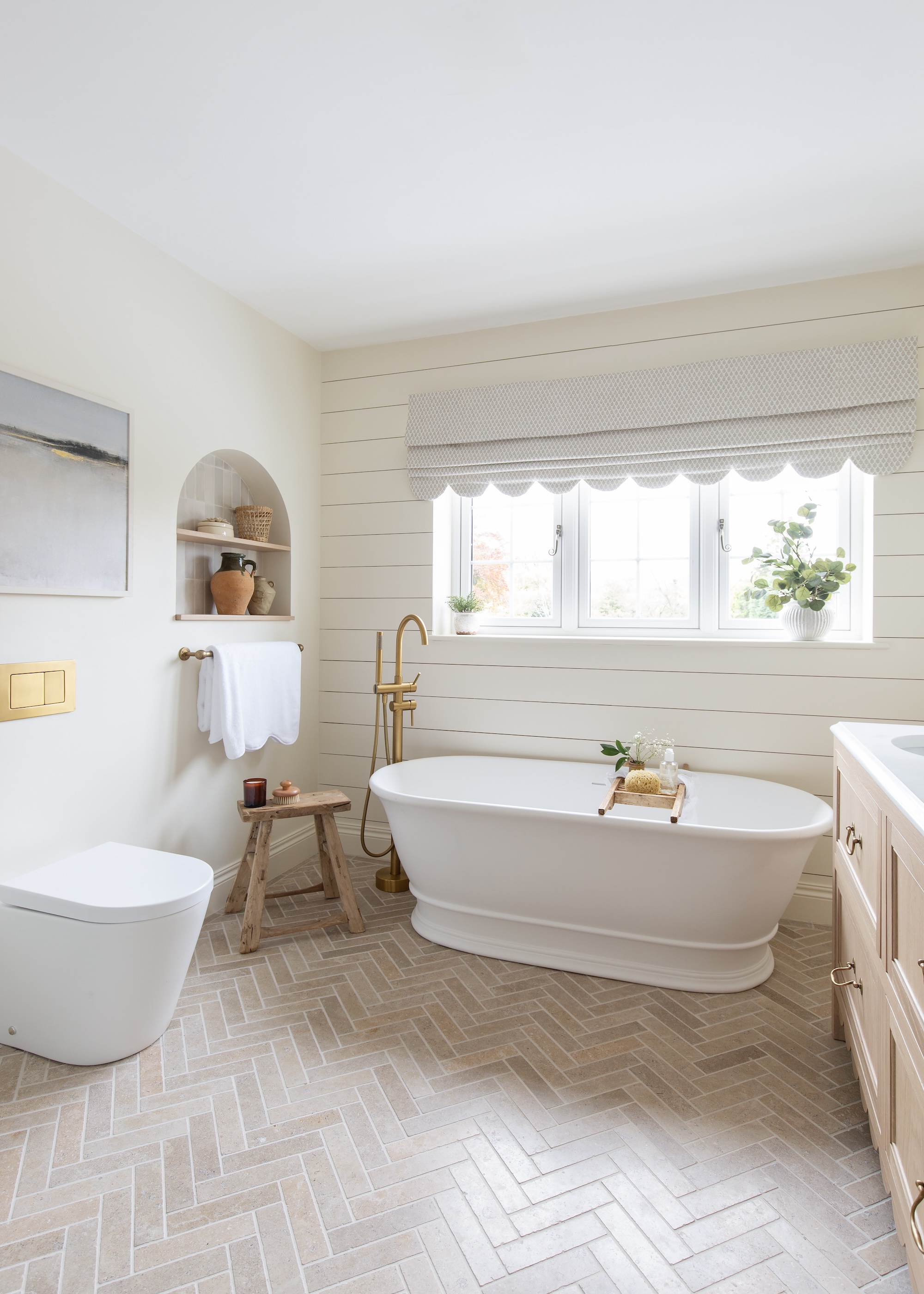 A white bathroom with herringbone floor tiles. There is also a bathtub and toilet in the space with gold features and hardware
