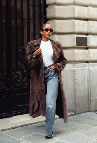 a photo of a woman walking on the sidewalk wearing a dark brown suede trench coat over a white t-shirt with jeans and pointed brown heels