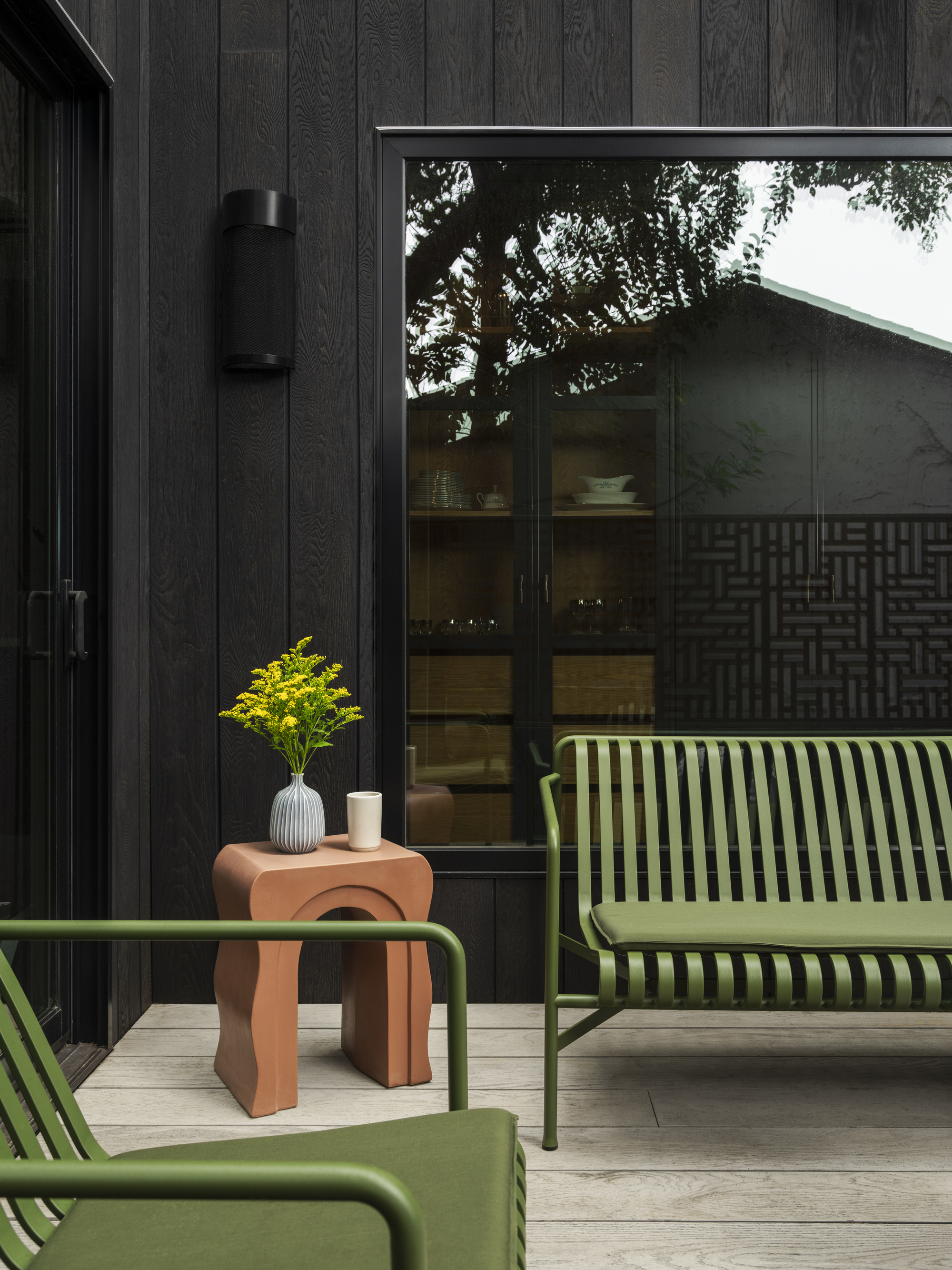 Image of a porch on the exterior of a black wooden house. There is green outdoor furniture and a terracotta accent table.