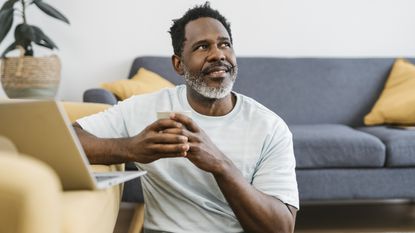 A smiling older man looks into the distance while sitting on the floor in front of his laptop on a coffee table.