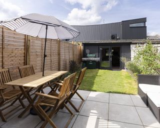 An external shot of backyard with garden outbuilding, wooden garden table and chairs and parasol with striped decor