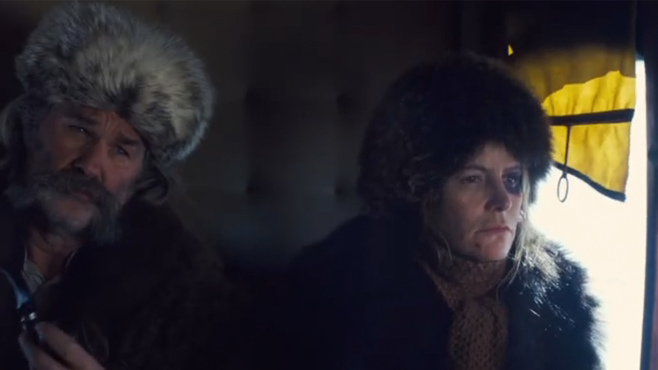 Kurt Russell in a big fur hat sitting next to Jennifer Jason Leigh, also in a fur hat in The Hateful Eight