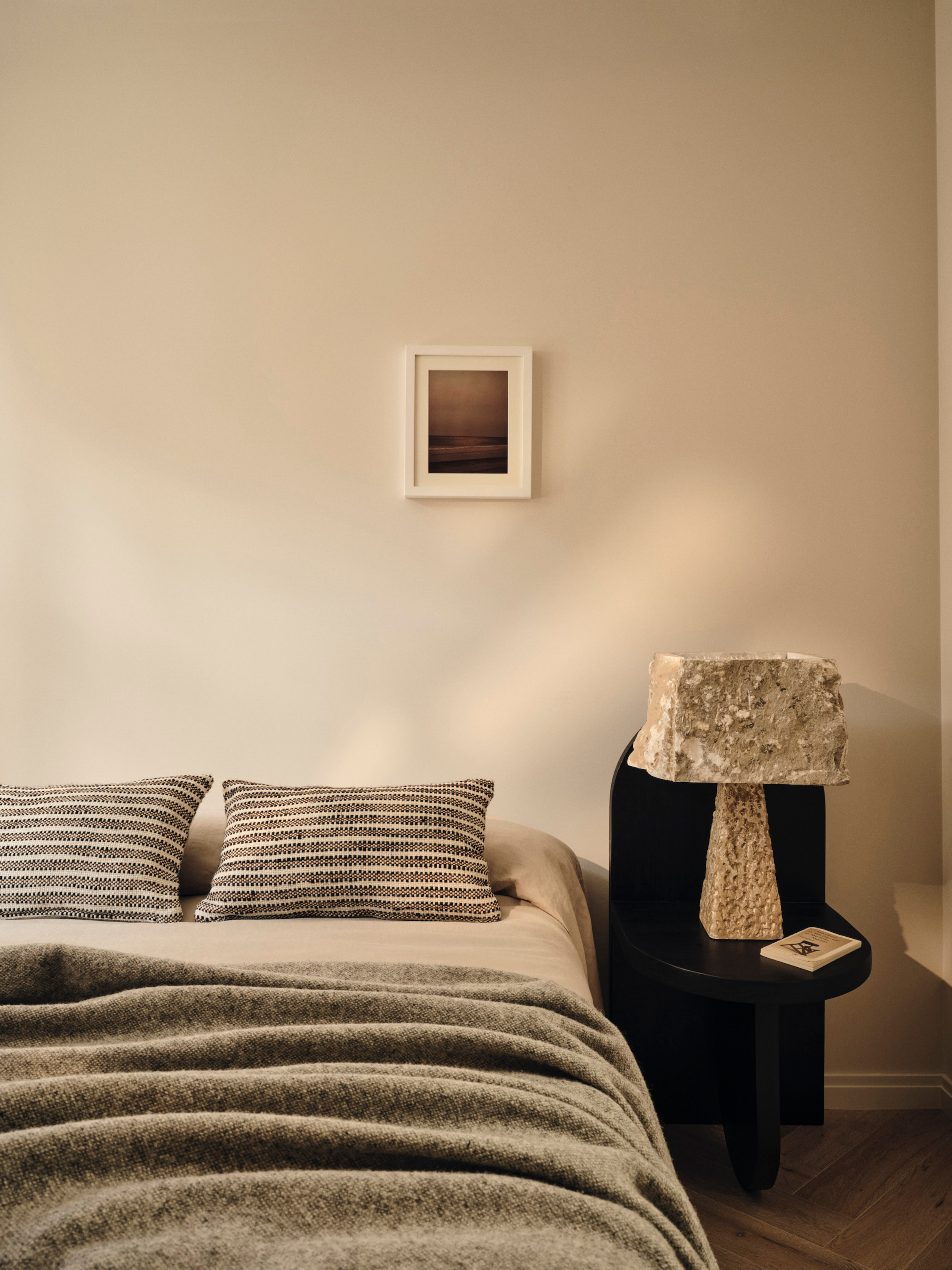 Bedroom with warm white walls, grey throw, striped grey and white cushions and black chair acting as bedside table with hammered effect bedside light
