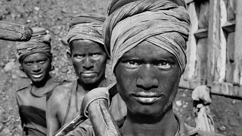 A close-up, black-and-white photograph shows three coal miners with dark, coal-dust covered faces and head wraps, with the man in the foreground looking directly at the camera while holding a shovel over his shoulder.