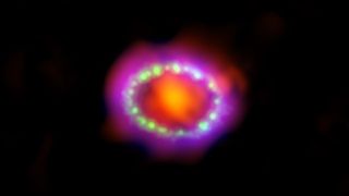 An image of SN 1987A, the brightest supernova seen in over 400 years. Here we see an orange-red circular center surrounded by a ring of white dots and then a purple and red ring.