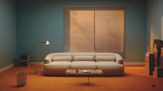 A living room with light teal walls and a bright orange rug. There is a window with blinds on the back wall and a white modern sofa in the middle of the room. A glass coffee table is in front of a sofa and there is a floor lamp, metal waste basket, and a stack of boxes also in the room.