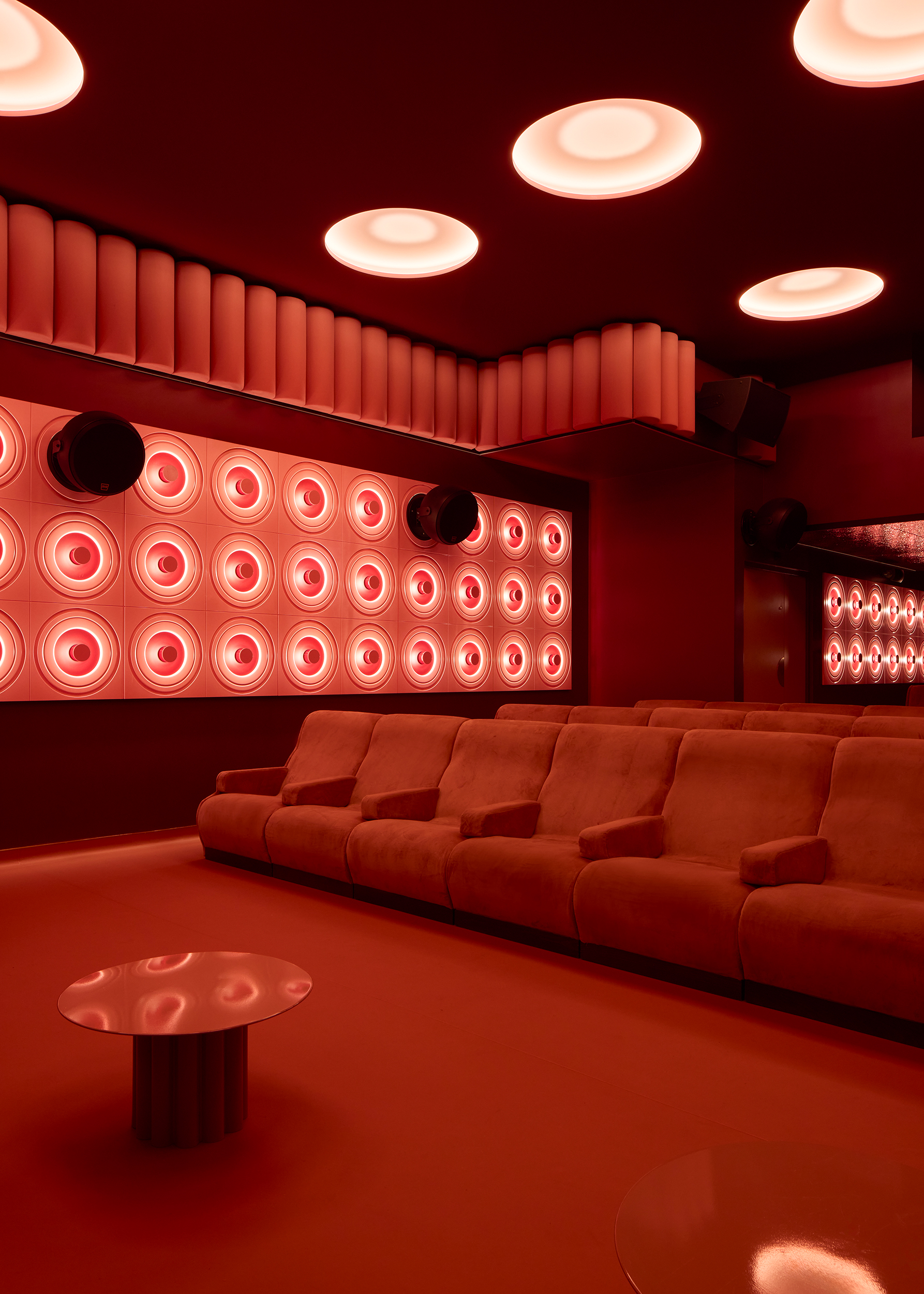 A cinema screening room with velvety red seats, porthole window-like ceiling lights, high-tech sound panels in pink, rounded shapes, and semi-circular portals.