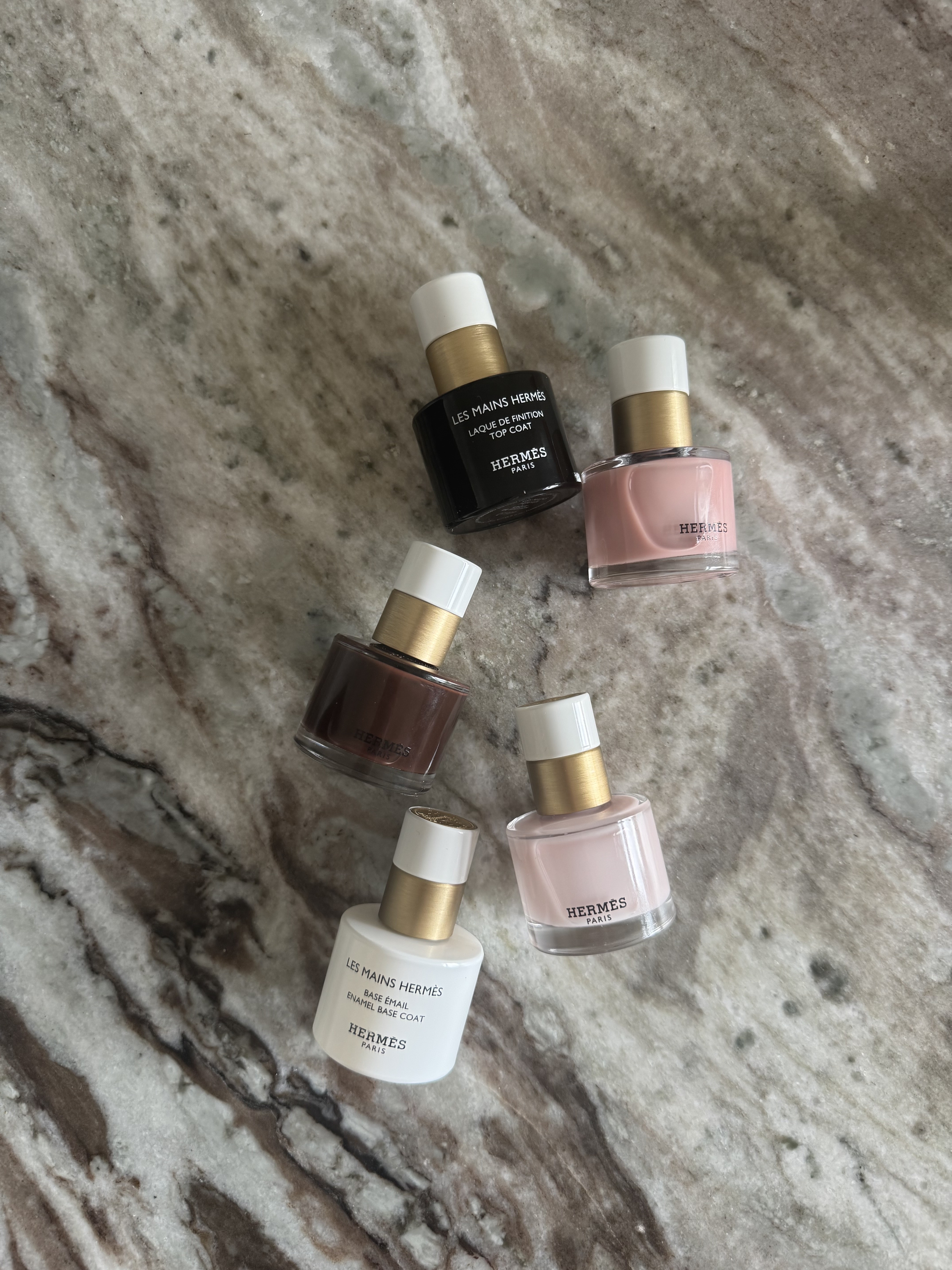Hermes nail polish bottles scattered on a marble textured background, including pink and brown shades alongside the base and top coats