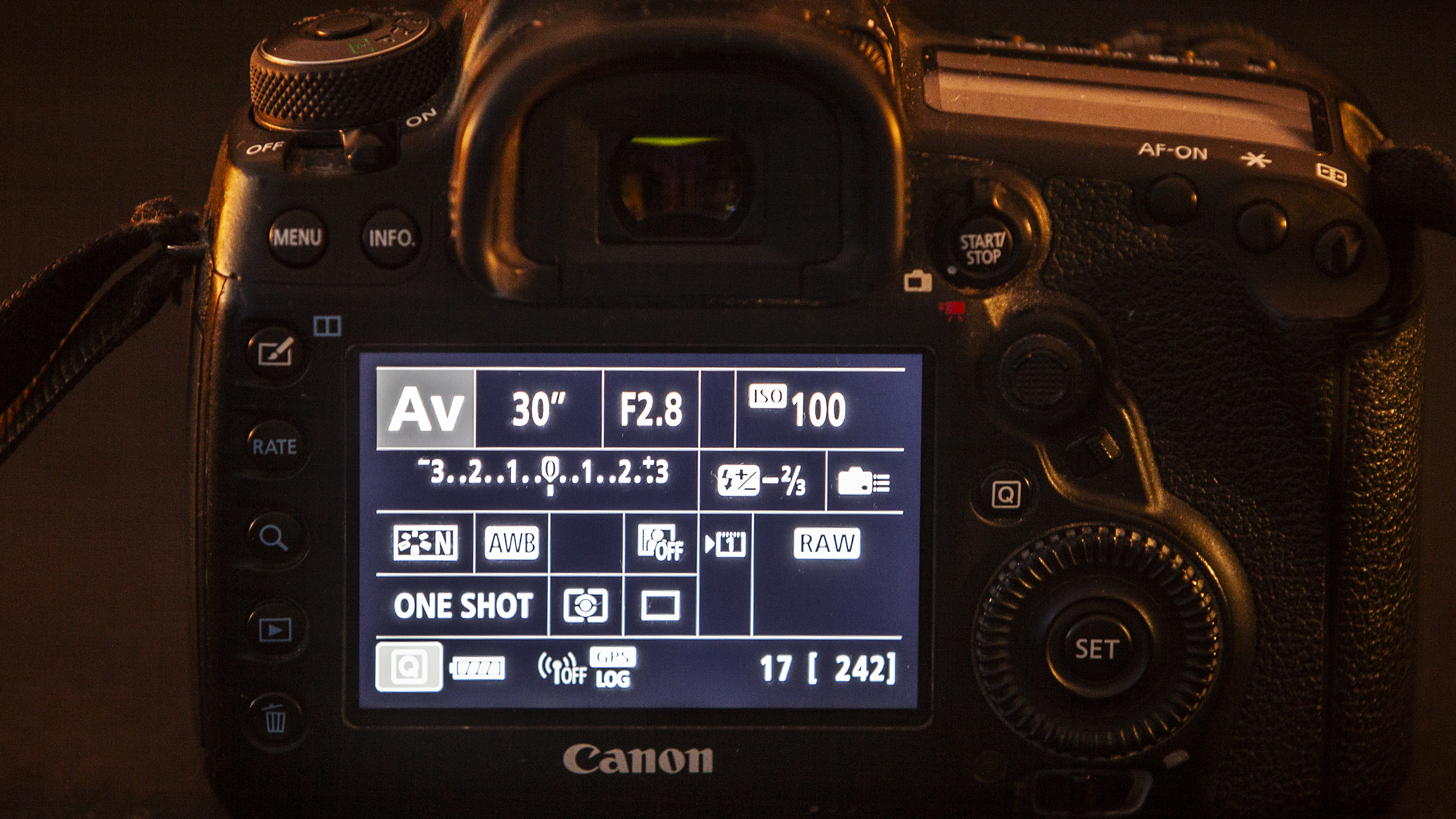 Close-up of a Canon DSLR back, focusing on the illuminated display. Settings shown include aperture, shutter speed, and ISO. Dark background
