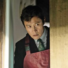 A man (Park Byung-hun as Man-su) wears an apron over a suit whole peeking through a doorway, in Park Chan-wook's 2025 movie 'No Other Choice.'