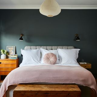 King size bed with white and pink bedding and grey headboard. In front of dark wall with wooden bedside table and ottoman