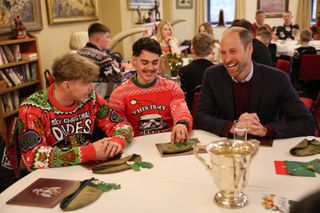 Prince William wears a burgundy sweater and suit jacket while talking to two men wearing bright red and green Christmas sweaters