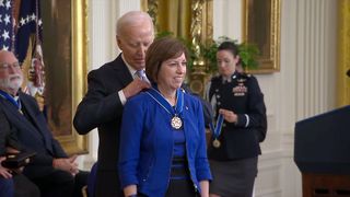 an old man wearing a suit drapes a medal around the neck of a smiling woman in a blue dress.