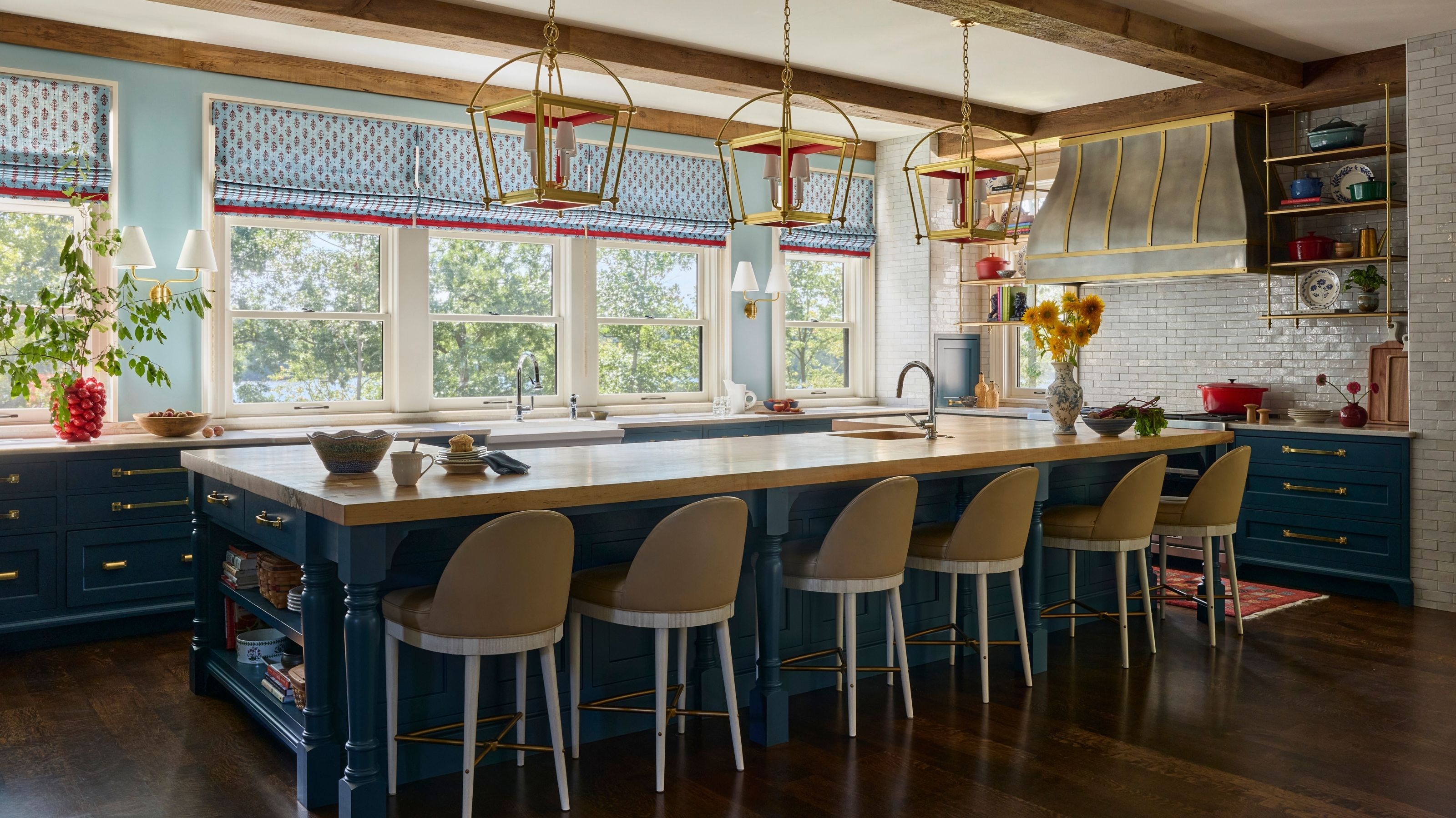 Spacious kitchen with blue cabinets, light blue walls, exposed ceiling beams, three brass lantern pendants, and a large wood-topped island lined with beige leather barstools.