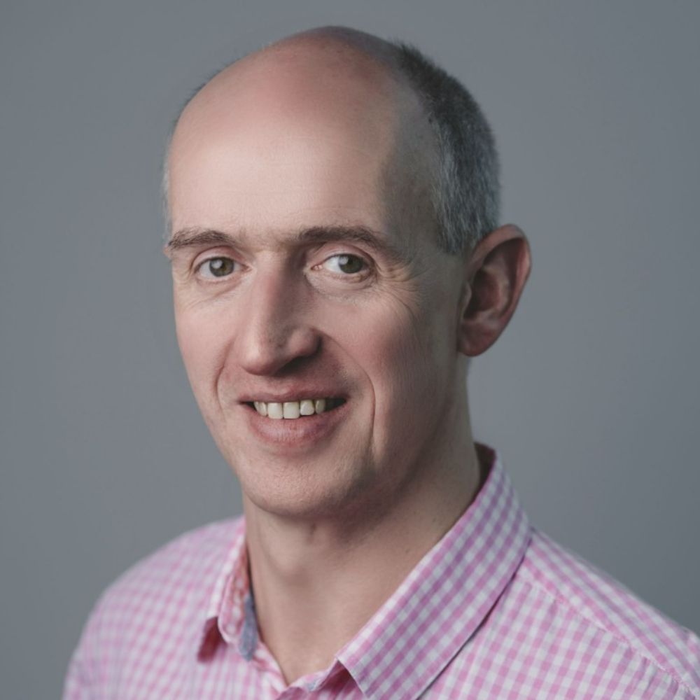 A smiling middle-aged man with close-cropped grey hair wearing a pink gingham shirt, photographed against a plain grey background.