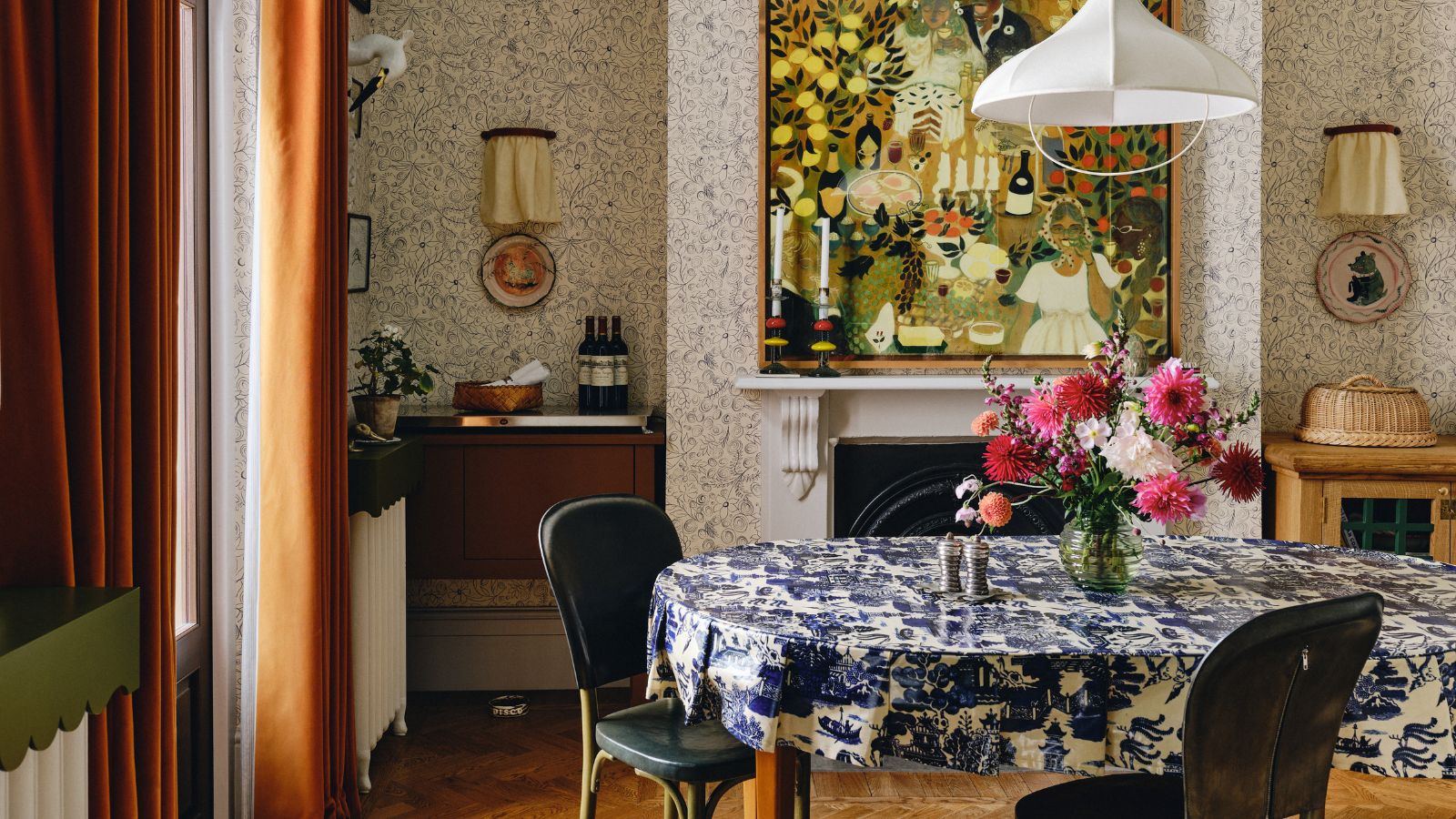 scandi maximalist style dining room with wallpapered walls, a large fireplace with folk art hung above, dark orange velvet curtains, a dining table with blue and white printed tablecloth and a white fabric pendant light