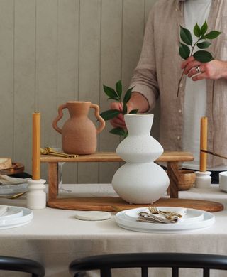 White, minimalist dining table with stacked white plates, orange candle sticks, and wooden food risers in the center. There are too organic shaped vases with a person putting green branches into them