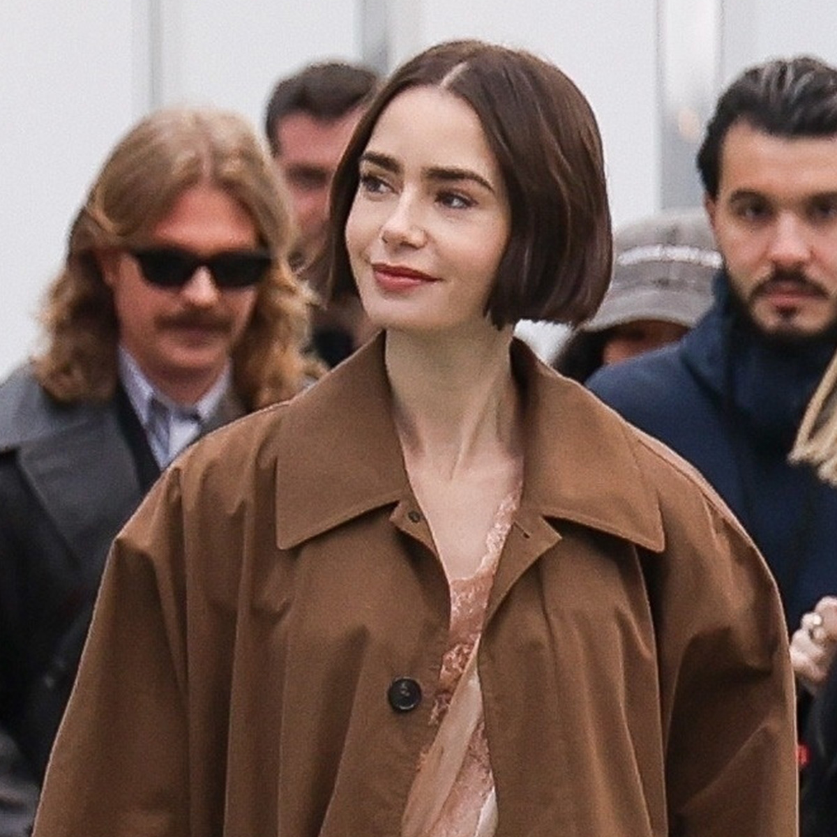 Lily Collins arrives at Gare d&rsquo;Austerlitz in Paris wearing a lingerie romper from YSL with a trench coat, tights, and heels, for Emily in Paris.