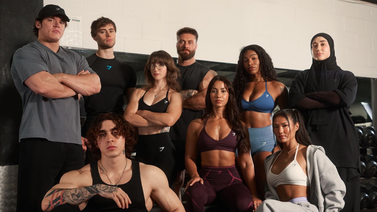 Nine Gymshark athletes pose wearing the brand&#039;s apparel. Behind them we see a dumbbell rack.