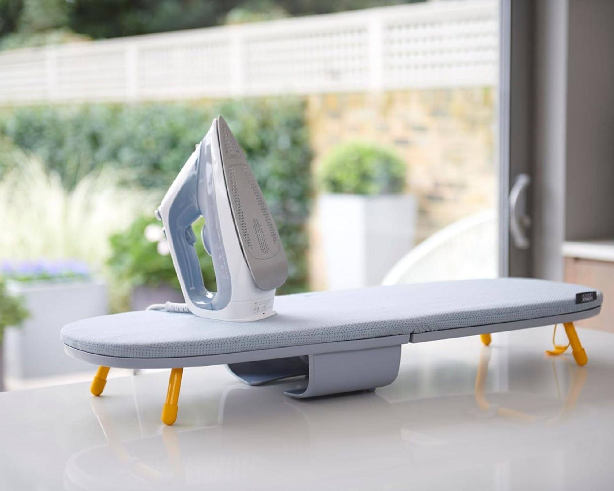 Joseph Joseph compact ironing board with blue iron on a white kitchen counter with a sunny garden seen through large windows behind